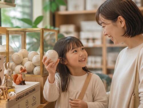 店内で紙素材のエコカプセルトイを手にして母親と笑顔で見つめ合う女の子と脱プラ商品のディスプレイ